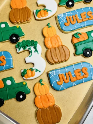 Cookies for a fall themed third birthday, pumpkins stacked on top of each other, pumpkin in a back of a truck, cookie with a spider web and the name "Jules", and a 3 with pumpkins, leaves, and vines