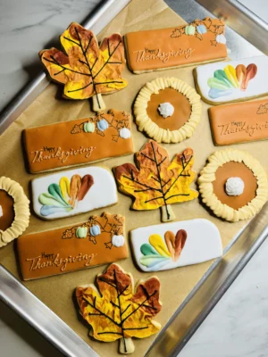 Thanksgiving cookies with a fall leaf, turkey feather cookie, pumpkin pie and pumpkin floral cookie with the words "Happy Thanksgiving".